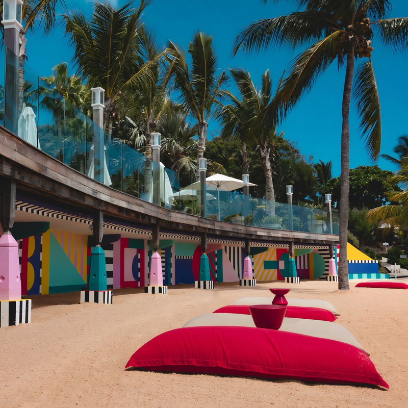 Coussin de plage rouge et blanc sur une plage de sable avec des palmiers avec des peintures multicolores sur un mur en arrière plan