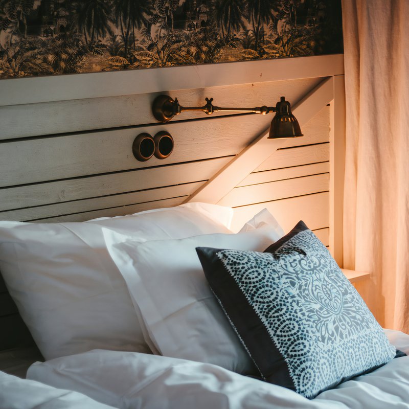 Lit avec une tête de lit en bois et une lampe de chevet murale et vintage. Trois coussins posés sur le lit