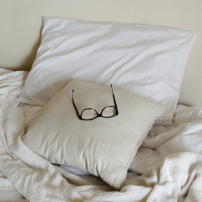 Housses de coussin beige et blanche sur un lit avec des lunettes posée sur le coussin beige